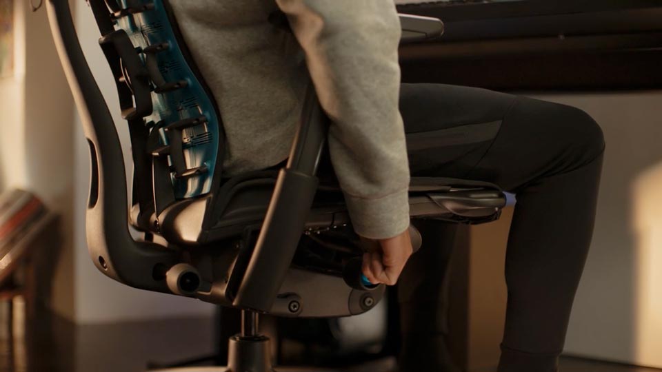 A video of a person sitting in a black and blue Embody gaming chair by Herman Miller, reaching beside the chair to adjust the seat height.