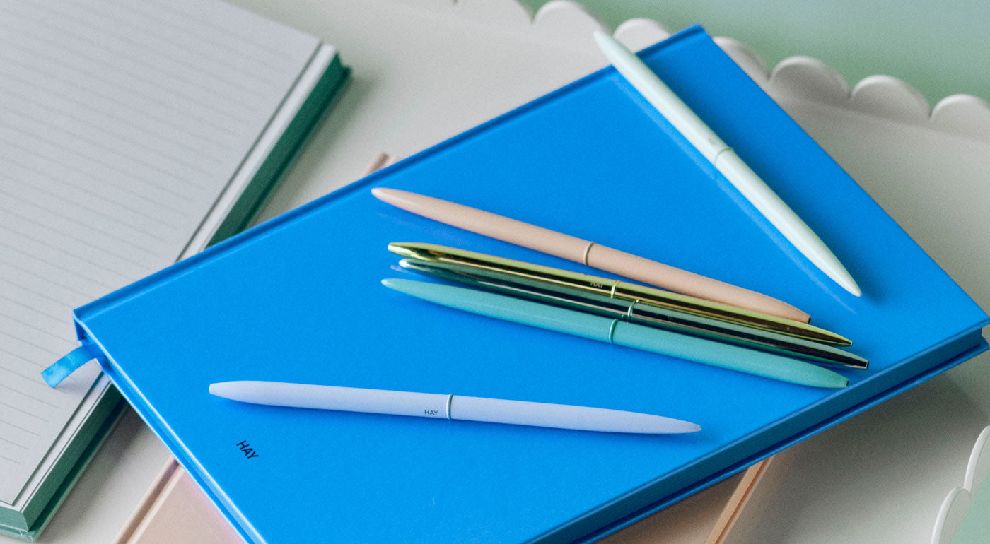 A selection of HAY Bullet Pens in various pastel and metallic colours sitting on a blue notepad.
