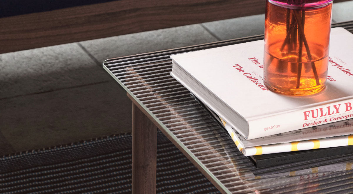 Close-up of a walnut Kofi coffee table by HAY, showing the reeded glass tabletop and styled with coffee table books and an orange glass vase.
