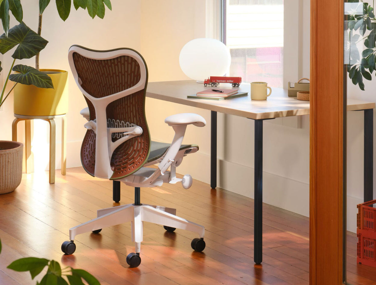 An olive green and white Mirra 2 office chair with a maroon back, positioned in front of a Herman Miller OE1 desk. Set in a home office environment, with solid wood flooring, white walls and warm, cosy lighting.