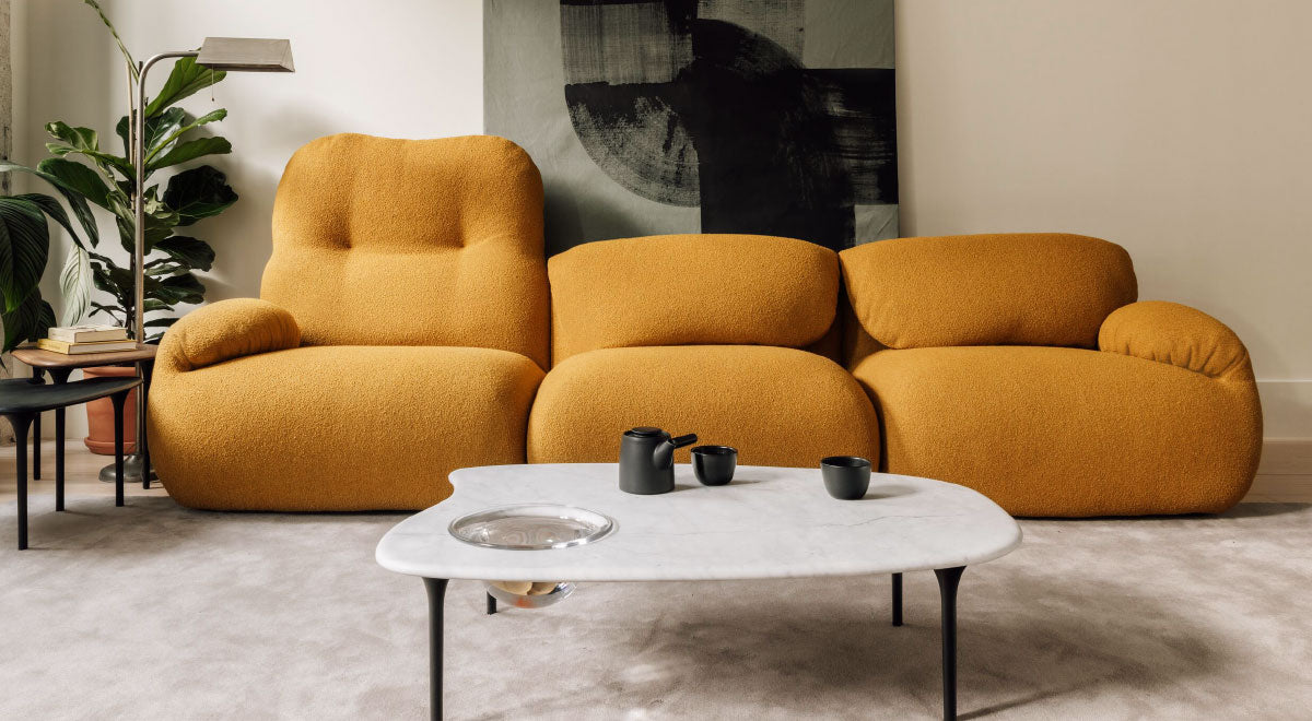 A mustard yellow 3-seater Luva sofa in Whim fabric with a marble top Cyclade coffee table featuring black metal legs. Various pieces of furniture, including a floor lamp, indoor plant and wall artwork complement the room's overall aesthetic.