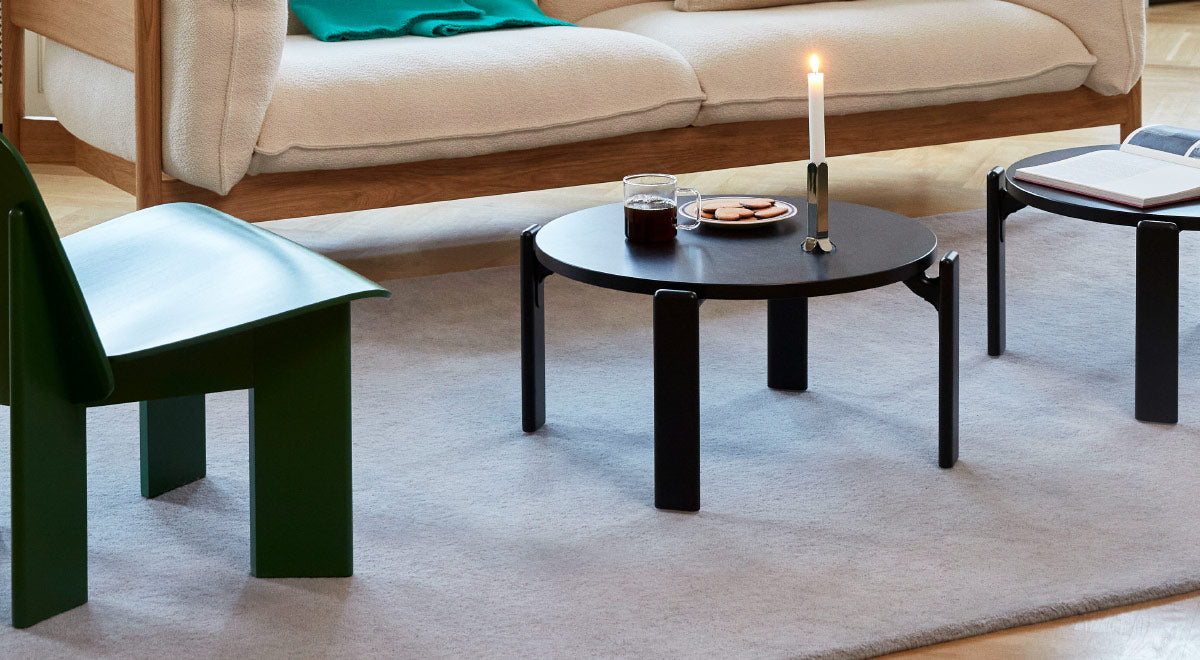 A black Rey coffee table by HAY in a cosy living room, featuring a Chisel lounge chair by HAY, a beige sofa and a grey rug. The coffee table is accessorised with a mug, plate and candlestick.