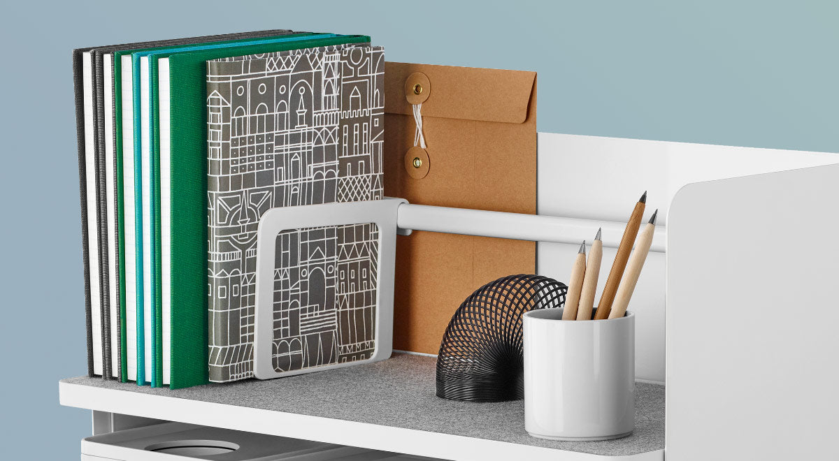 A white Ubi desktop organiser by Herman Miller, placed on the edge of a white desk. Featuring a grey shelf liner, white bookend and a white Formwork stationary box by Herman Miller.