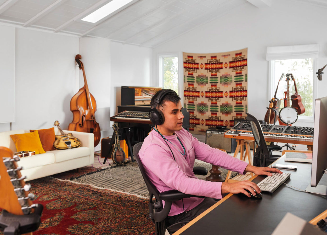 Rostam, musician, producer and composer, sat in Aeron office chair in graphite at computer with headphones on with musical instruments in background.