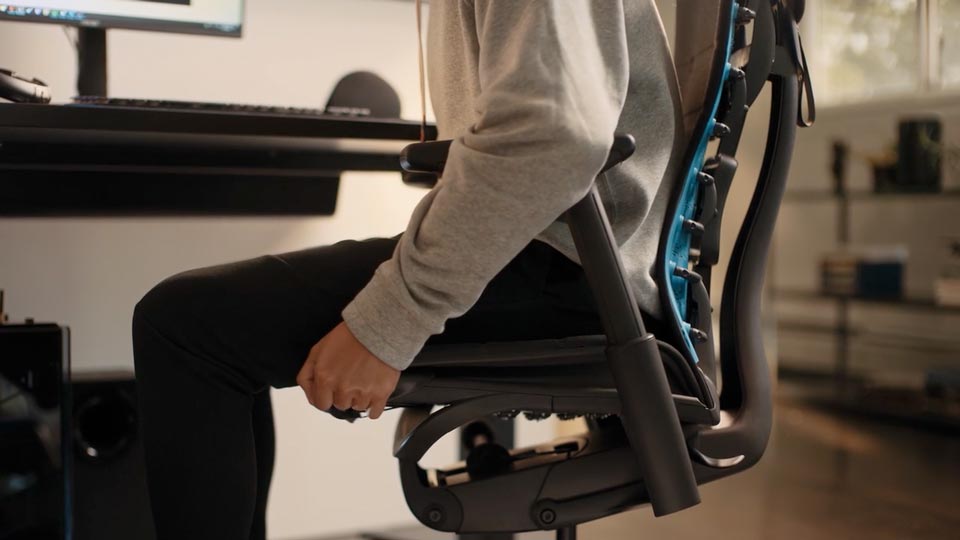A video of a person sitting in a black and blue Embody gaming chair by Herman Miller, reaching beside the chair to adjust the seat depth.