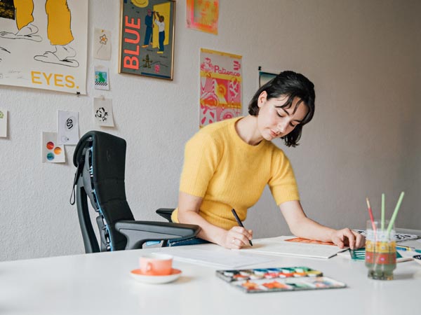 Illustrator, painter and author Molly Rose Dyson working in her studio workspace sat in an Embody gaming chair in black and cyan blue