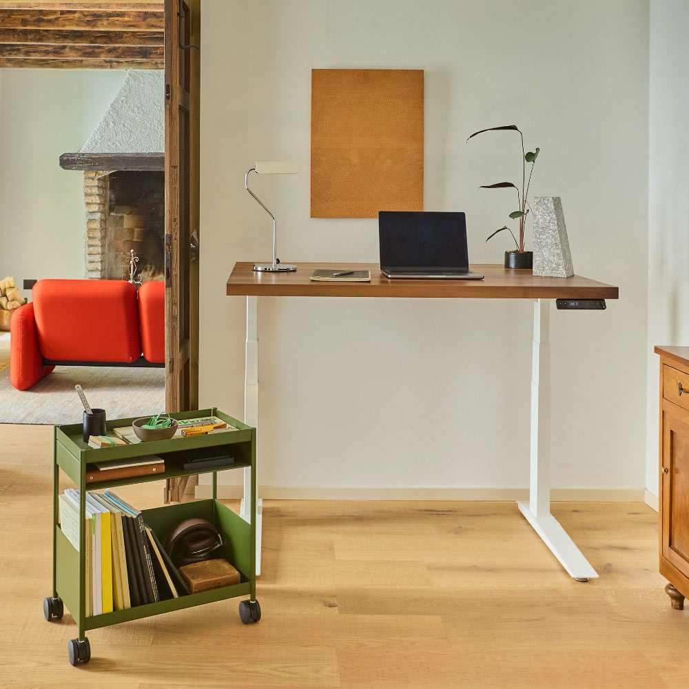 A Herman Miller Jarvis sit-stand desk in its highest position; featuring an olive green OE1 trolley, red Wilkes chiclet sofa and HAY Apex lamp.