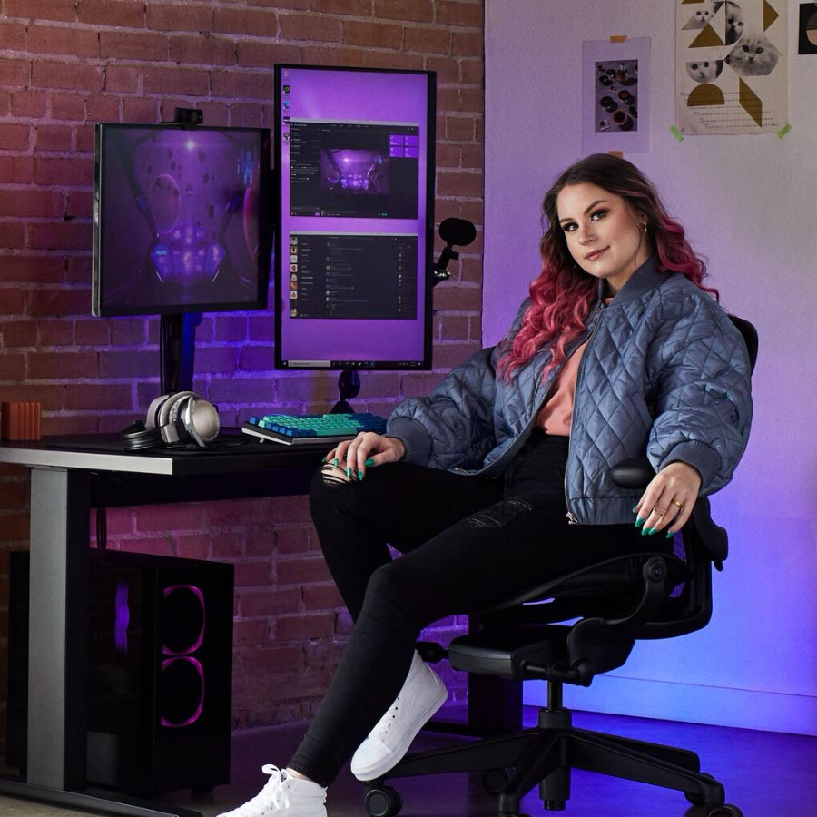 Star gamer and coach Electra sat at gaming set containing Herman Miller Aeron gaming chair with Nevi sit-stand desk in black with dual monitors on Ollin monitor arms