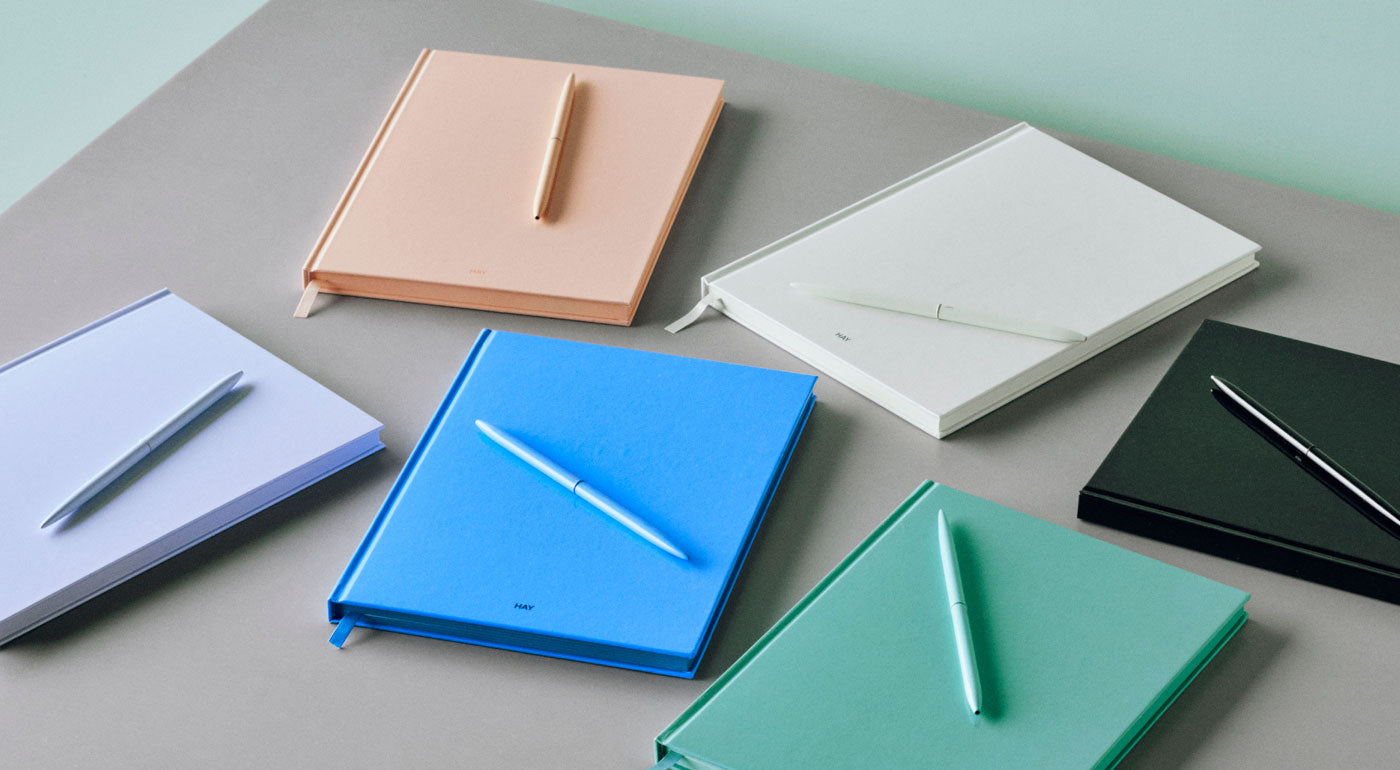 A selection of HAY Colour Notebooks in purple, peach, blue, white, green and black with colour-matching pens.