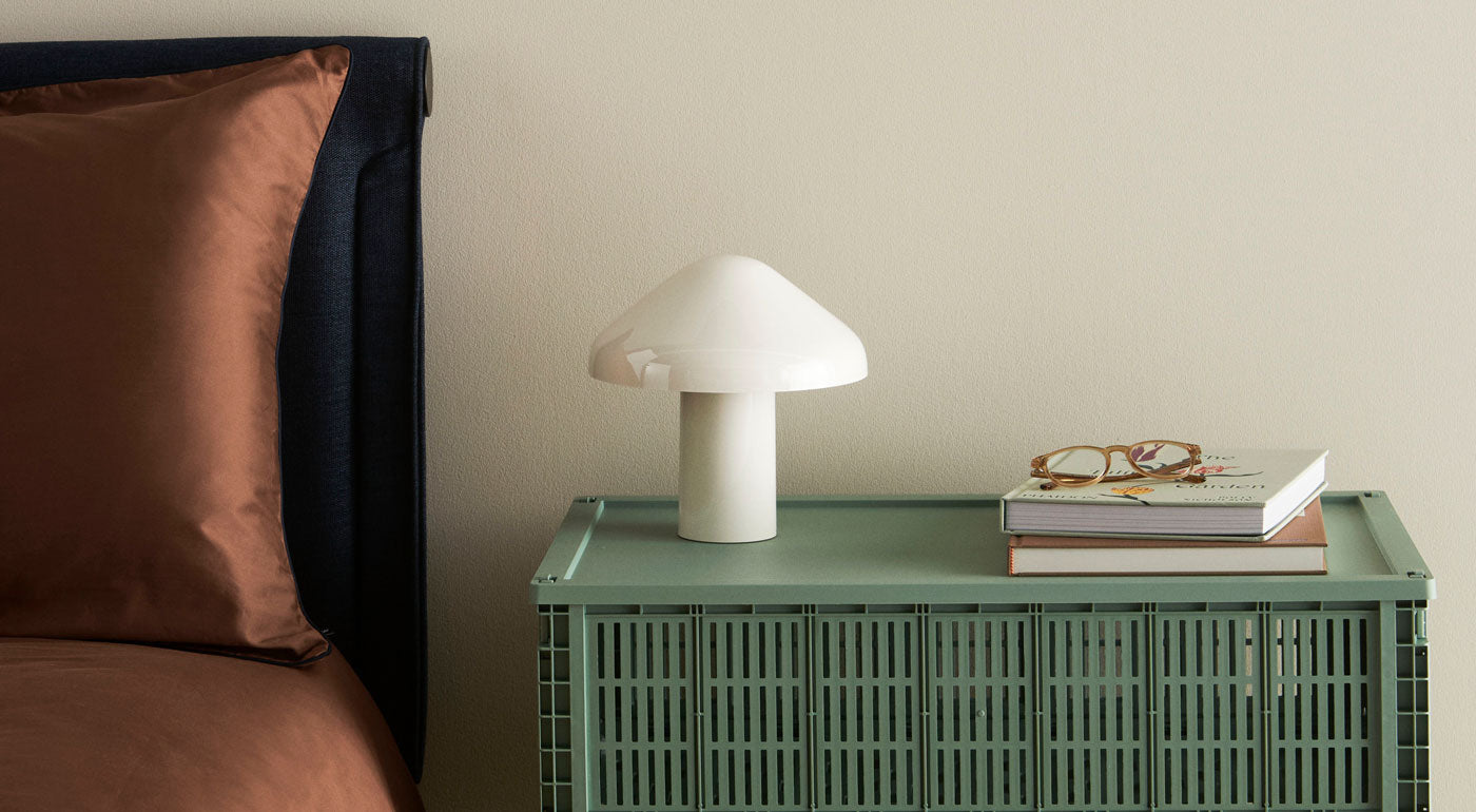 An Extra Large HAY Crate with a lid being used as a bedside table featuring a portable lamp, books and glasses.