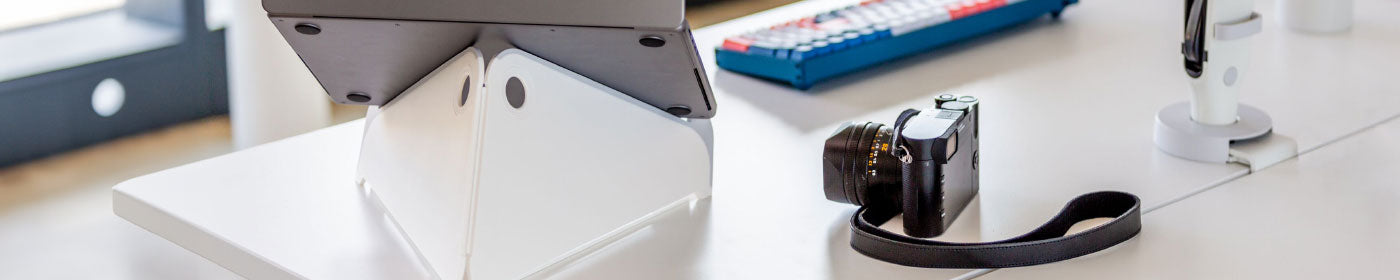 A white desk with a Macbook Pro sat on an Oripura laptop stand, compact mirrorless camera and an Ollin monitor arm.
