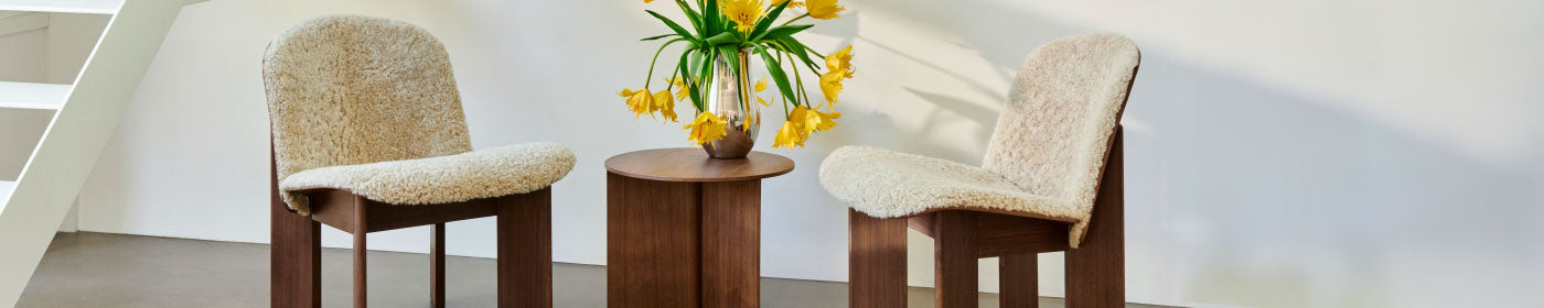 Two fleece-covered HAY Chisel lounge chairs in a dark walnut finish with a Slit side table and a vase containing daffodils.
