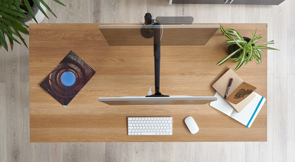 Top view of a black Lima monitor arm by CBS attached to a wooden desk, showing the movement of the monitor arm and screen.