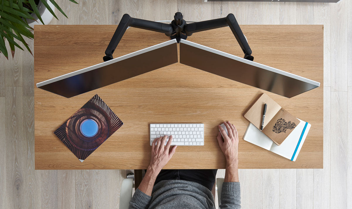 Top view of a wooden desk with a dual Lima monitor arm setup holding two screens.