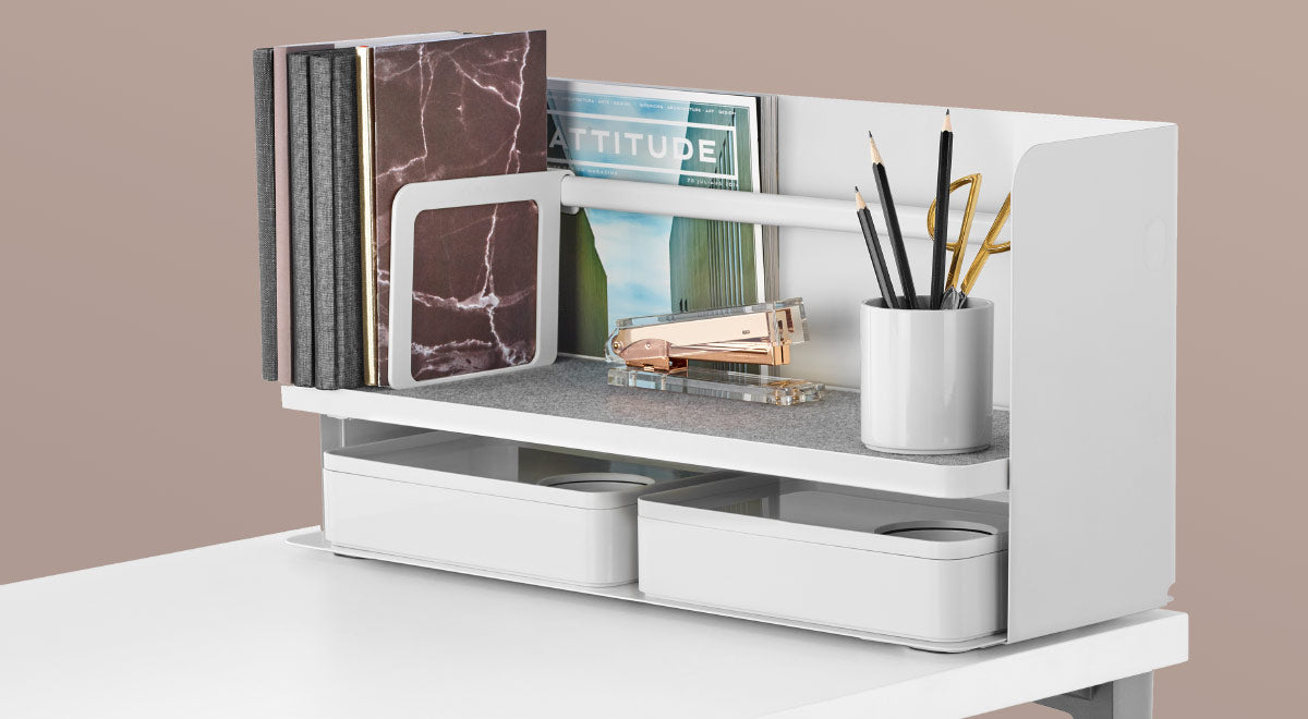 A white Ubi desktop organiser by Herman Miller, placed on the edge of a white desk. Featuring a grey shelf liner, white bookend and two white Formwork stationary boxes by Herman Miller.